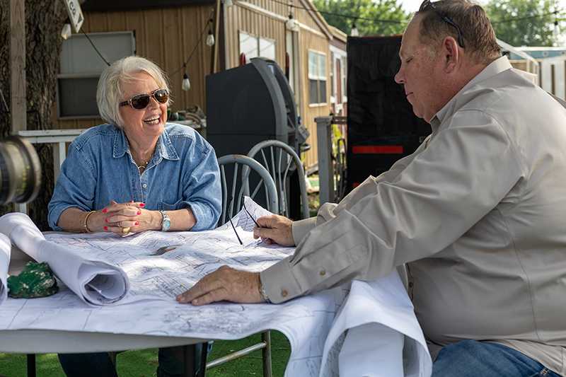 Woman in denim shirt and black sunglasses talking with a man in a tan shirt as they look at drilling plans spread out on a table